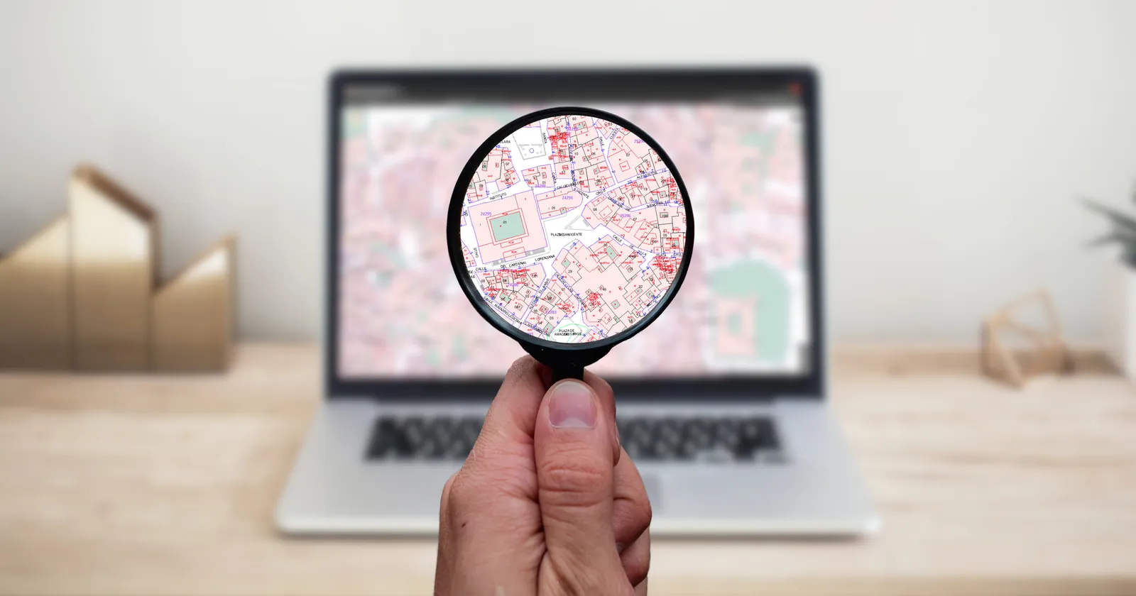 A hand holds a magnifying glass up to a laptop screen, enlarging part of a street map while the rest of the desk stays out of focus.